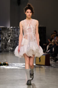 Female model walks the runway in a sheer white lace dress with floral appliqués, paired with gray socks and sneakers, at a fashion show audience in background.