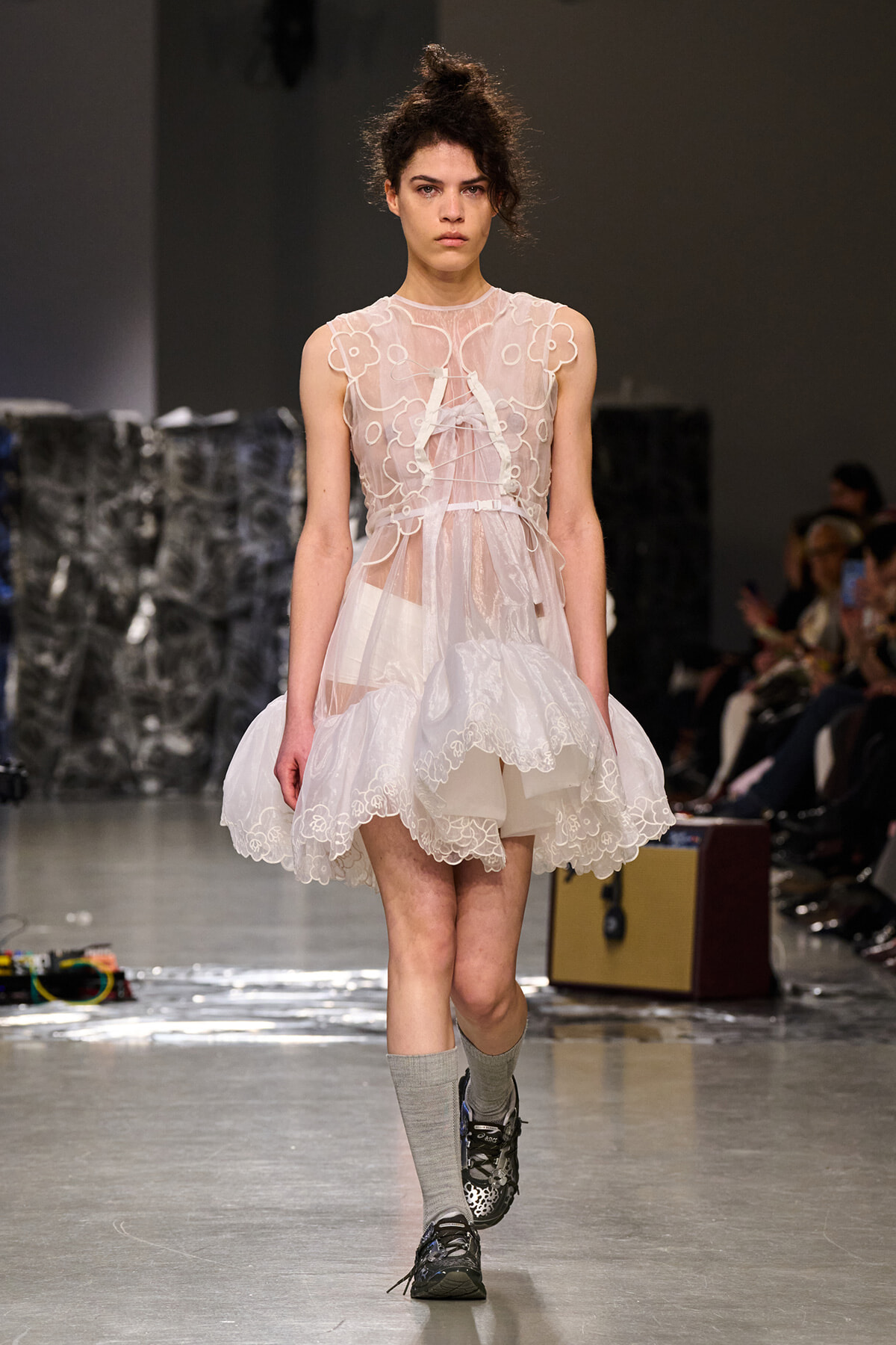 Female model walks the runway in a sheer white lace dress with floral appliqués, paired with gray socks and sneakers, at a fashion show audience in background.