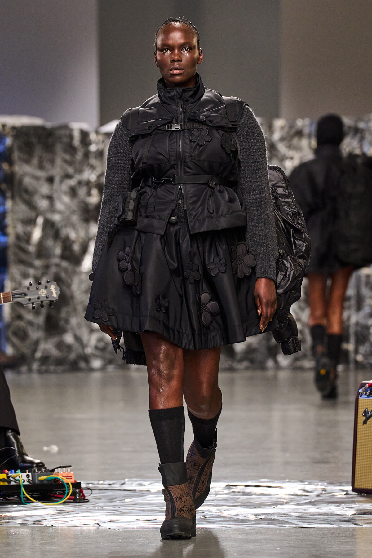 Female model walking a runway in a black padded dress with floral appliqués, wearing a backpack and boots.