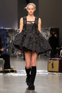 Model walking a runway in a dark grey, flower-embellished, voluminous dress with a structured bodice and black straps.