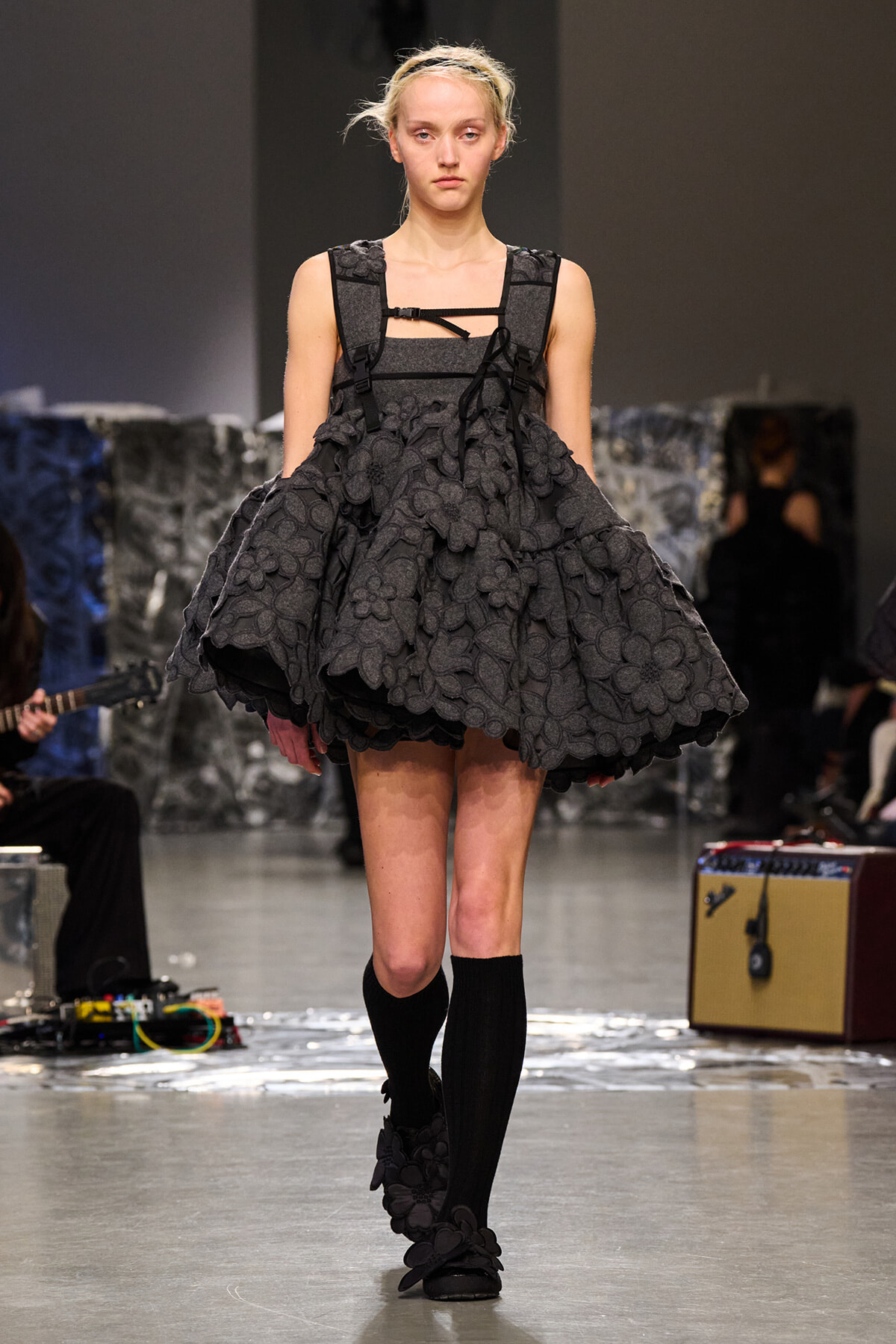 Model walking a runway in a dark grey, flower-embellished, voluminous dress with a structured bodice and black straps.
