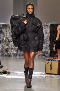 Model on a runway wearing a black hooded puffer coat with floral appliqués and a large backpack slung over one shoulder.