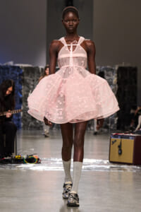 Black model walking a runway in a pale pink sheer dress with floral appliqués and ribbon straps.