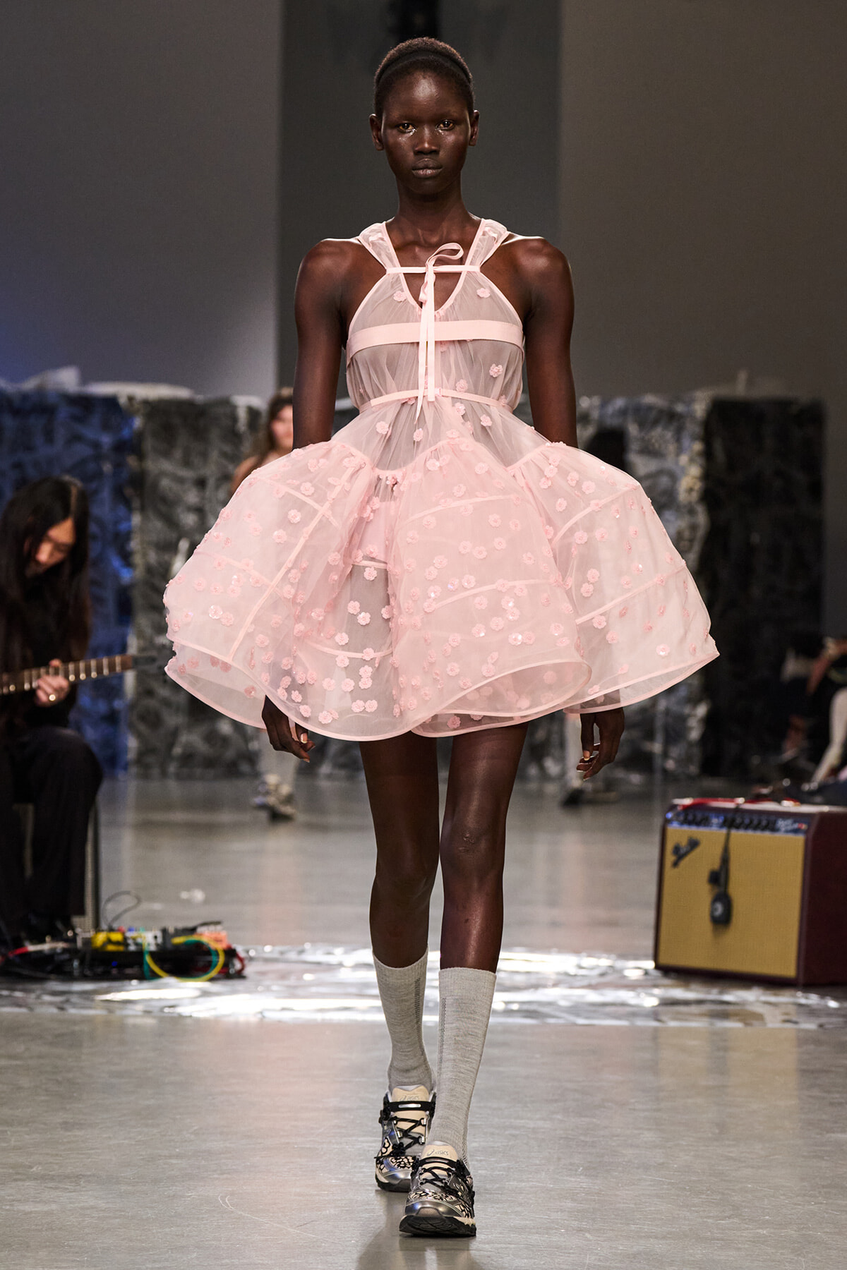 Black model walking a runway in a pale pink sheer dress with floral appliqués and ribbon straps.