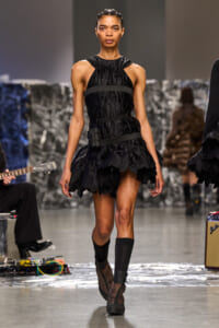 Model walks a fashion runway in a black quilted mini dress with layered ruffles and wide waist straps, paired with lace boots and a headband.