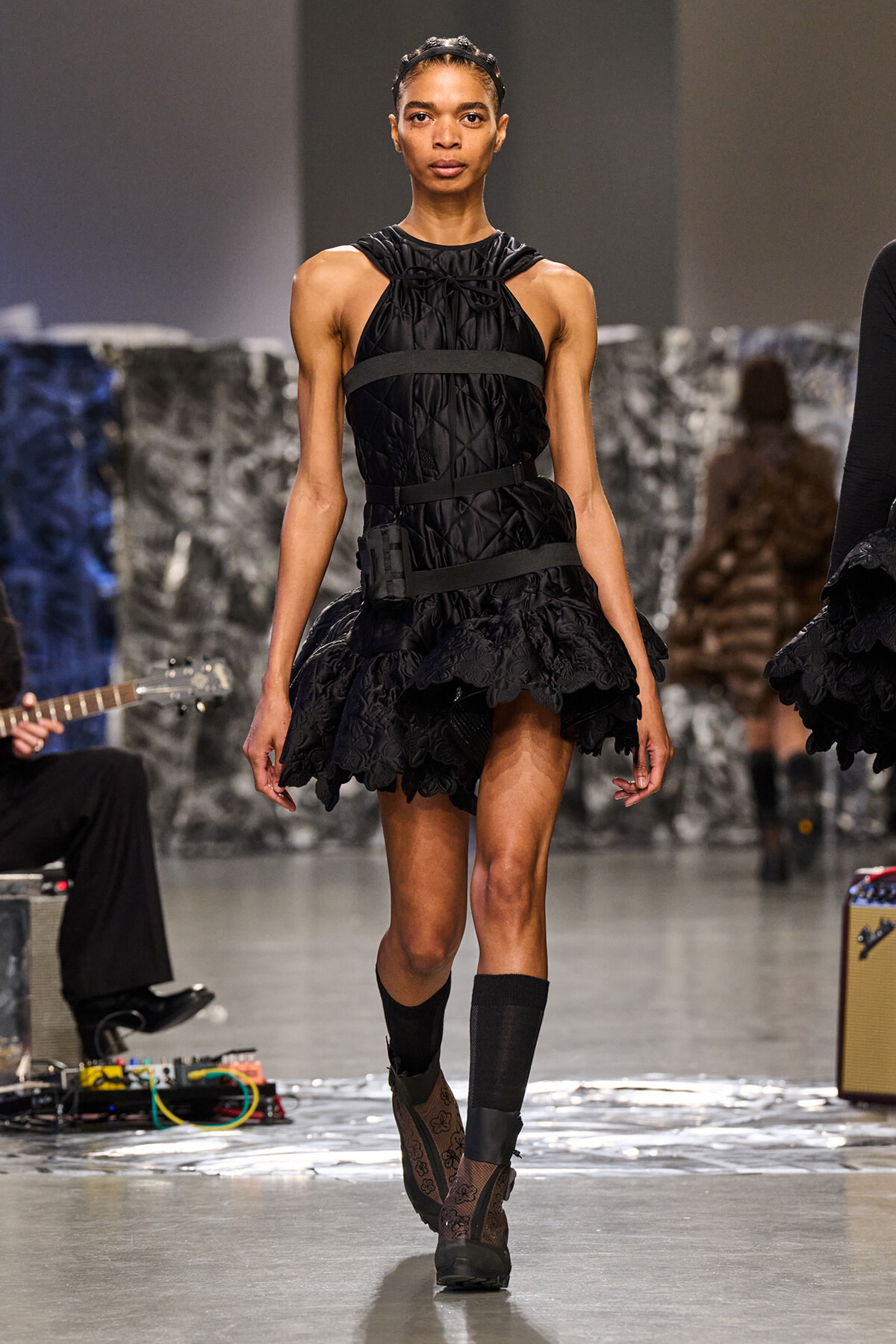 Model walks a fashion runway in a black quilted mini dress with layered ruffles and wide waist straps, paired with lace boots and a headband.