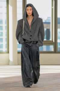 Person wearing an oversized gray hooded knit cardigan with a black belt and wide-leg dark jeans, indoors with large windows behind.