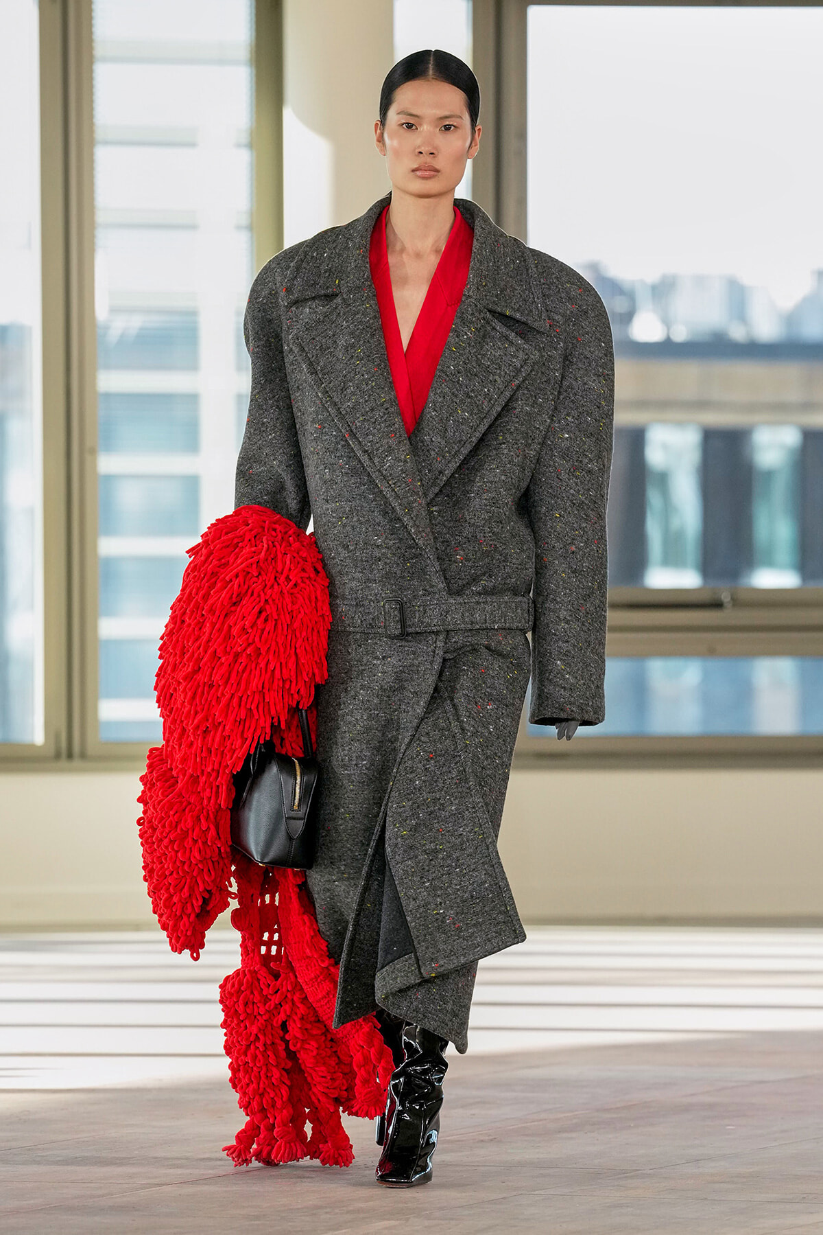 Model wearing a gray textured oversized coat with a red blouse, holding a bright red shaggy scarf and a black handbag against an urban backdrop.