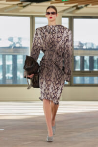 Model walking a runway in a gray snakeskin-print dress with a black chain-strap clutch and dark sunglasses.