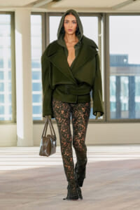 Model wearing an olive-green oversized coat with a hood, belted waist, and black lace leggings, carrying a gray handbag indoors.