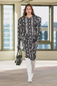 Model walking indoors in a black-gray geometric-print dress, white knee-high patent boots, gray gloves, and a black handbag.