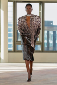 Model walking a runway wearing a purple snakeskin blouse with a deep neckline and a black metallic midi skirt indoors.
