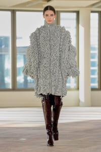 Model in a voluminous light-gray boucle coat with chunky looping texture and a high collar, paired with burgundy patent knee-high boots in a sunlit studio setting.