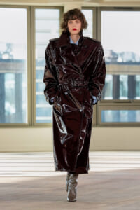 Model in a glossy burgundy trench coat with a belt, standing indoors by large windows.