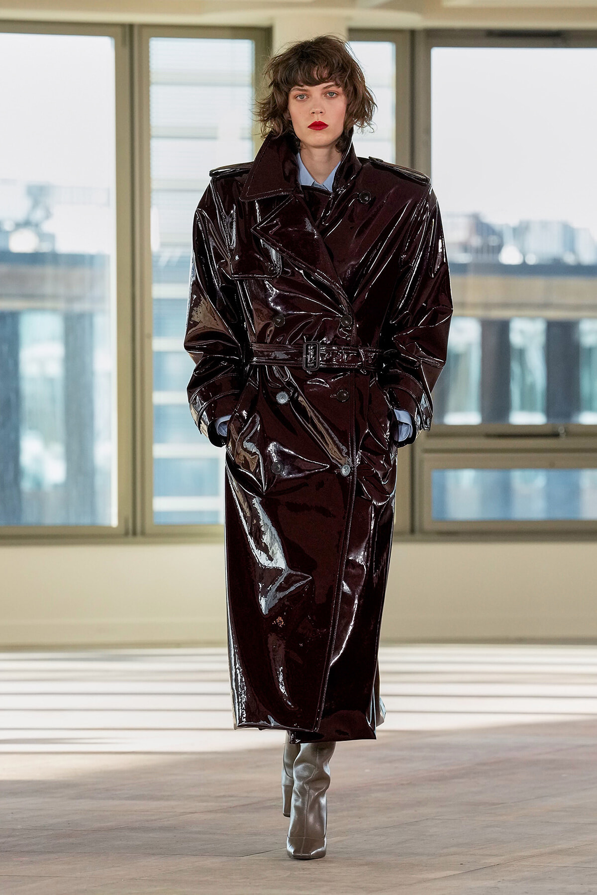Model in a glossy burgundy trench coat with a belt, standing indoors by large windows.