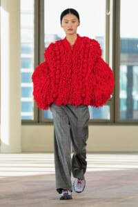 Model walks the runway wearing a voluminous red chunky-knit cape over gray tailored trousers in a bright indoor space.