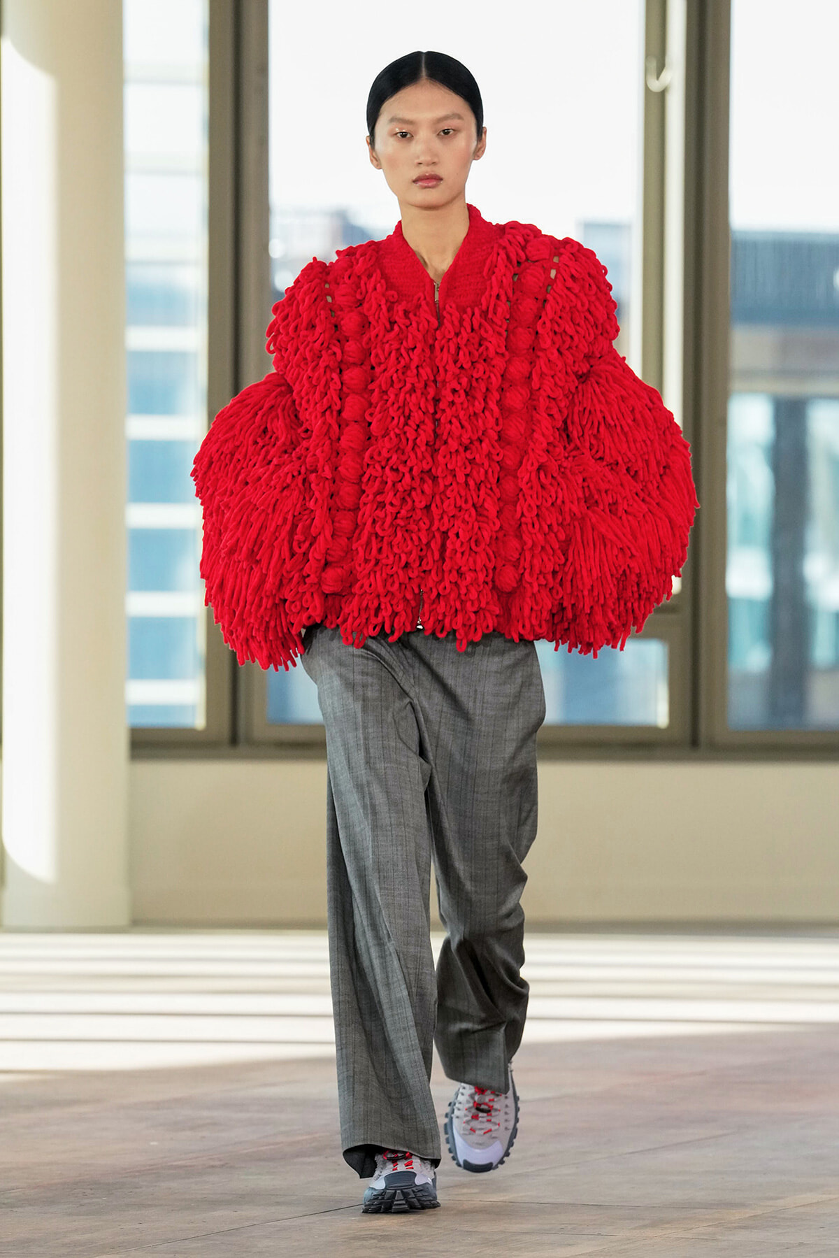 Model walks the runway wearing a voluminous red chunky-knit cape over gray tailored trousers in a bright indoor space.