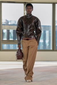 Model wearing a metallic snakeskin jacket with tan wide-leg trousers, carrying a burgundy handbag indoors.