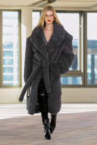 Model walking in a fashion setting wearing a long dark gray faux-fur coat tied with a belt, black patent boots, and a gray handbag.