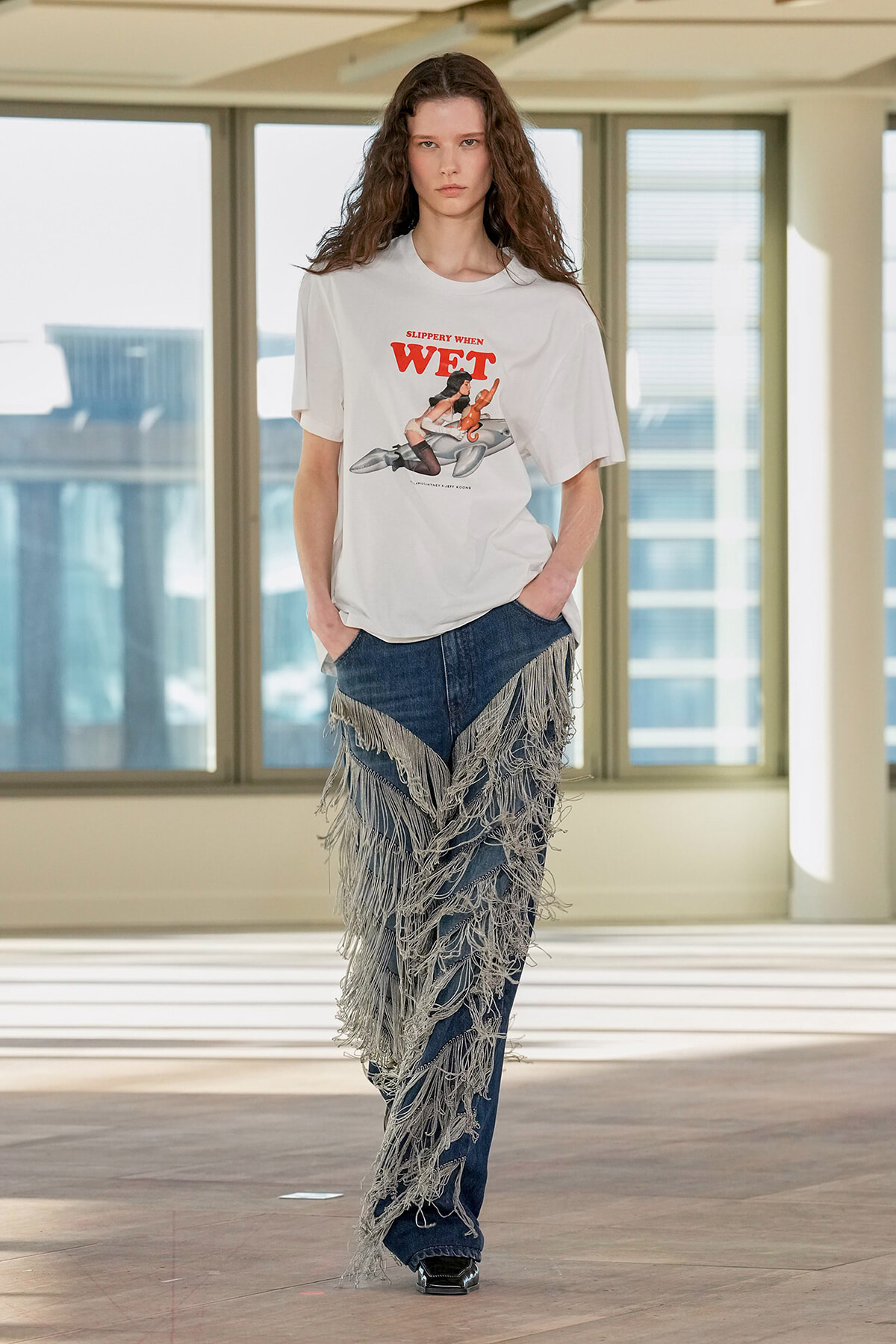 Female model wearing a white graphic T-shirt with a red 'WET' print, paired with blue denim pants featuring long fringe details, indoors in a bright studio.