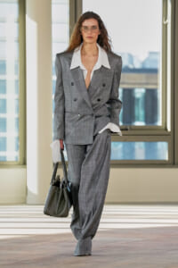 Woman in a gray double-breasted suit with an oversized white shirt, wearing glasses and carrying a black handbag, walking indoors by large windows.