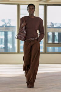Full-body shot of a person in a brown monochrome outfit walking indoors, carrying a textured brown shoulder bag.