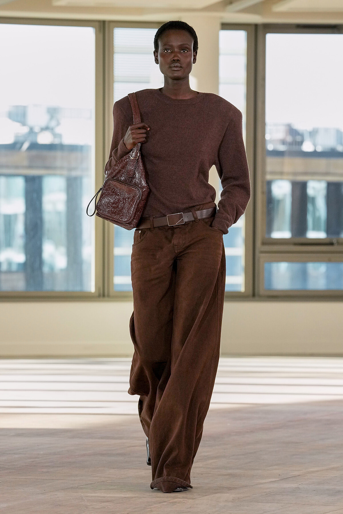 Full-body shot of a person in a brown monochrome outfit walking indoors, carrying a textured brown shoulder bag.
