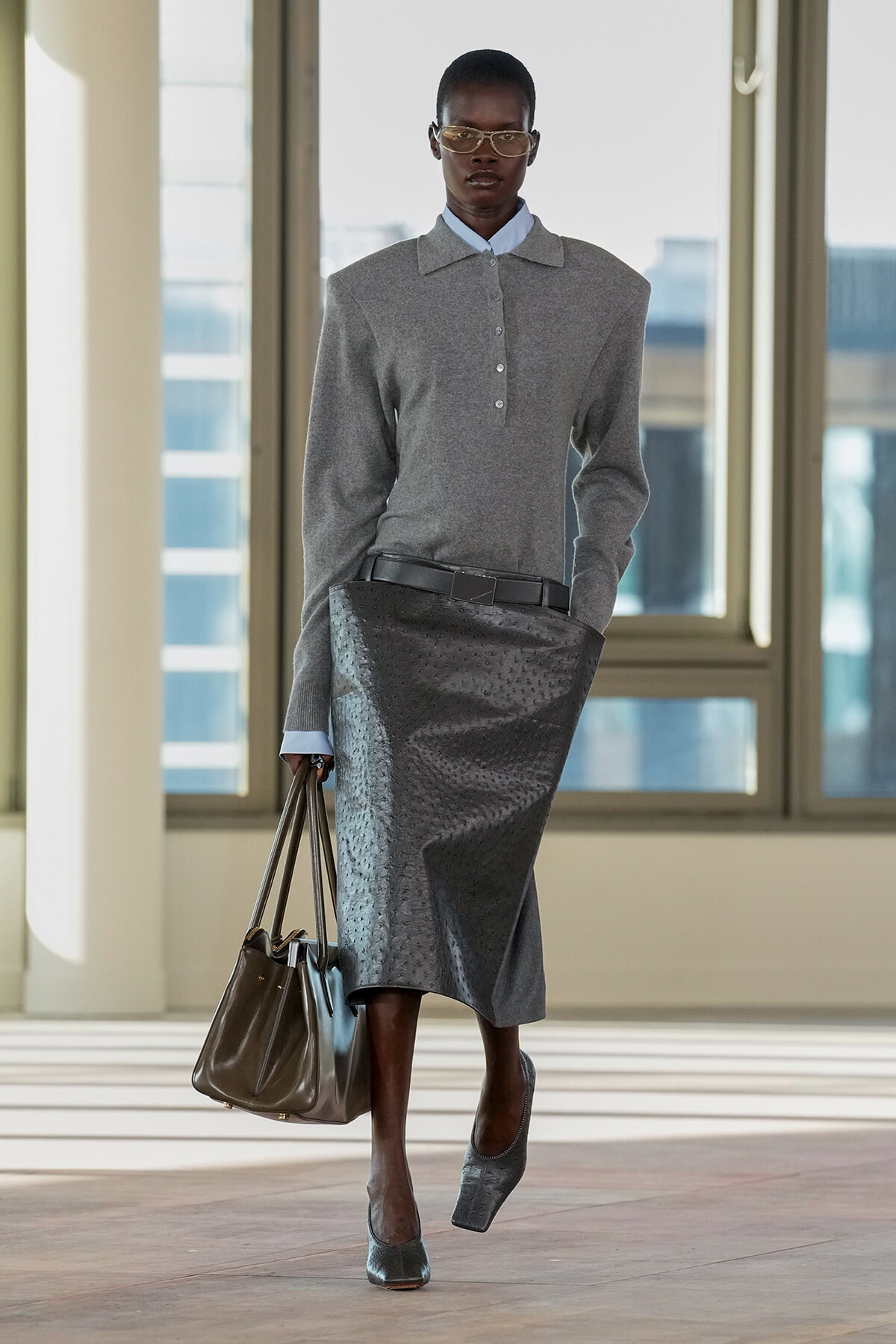 Model in a gray polo-knit top with a textured midi skirt, belt, gray heels, glasses, and a brown handbag walking in a bright atrium.
