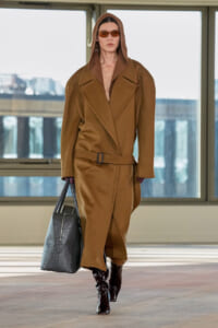 Model wearing a camel oversized coat with hood, belt at the waist, and a black textured tote bag.