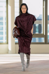 Model walks in a burgundy knot-front dress with a twisted waist, carrying a shiny brown clutch, wearing grey patent knee-high boots in a bright indoor space buttressed by windows.