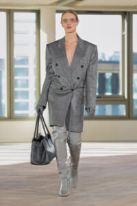 Model walking in a gray double-breasted blazer, shiny gray knee-high boots, and a textured gray tote bag in an urban interior.