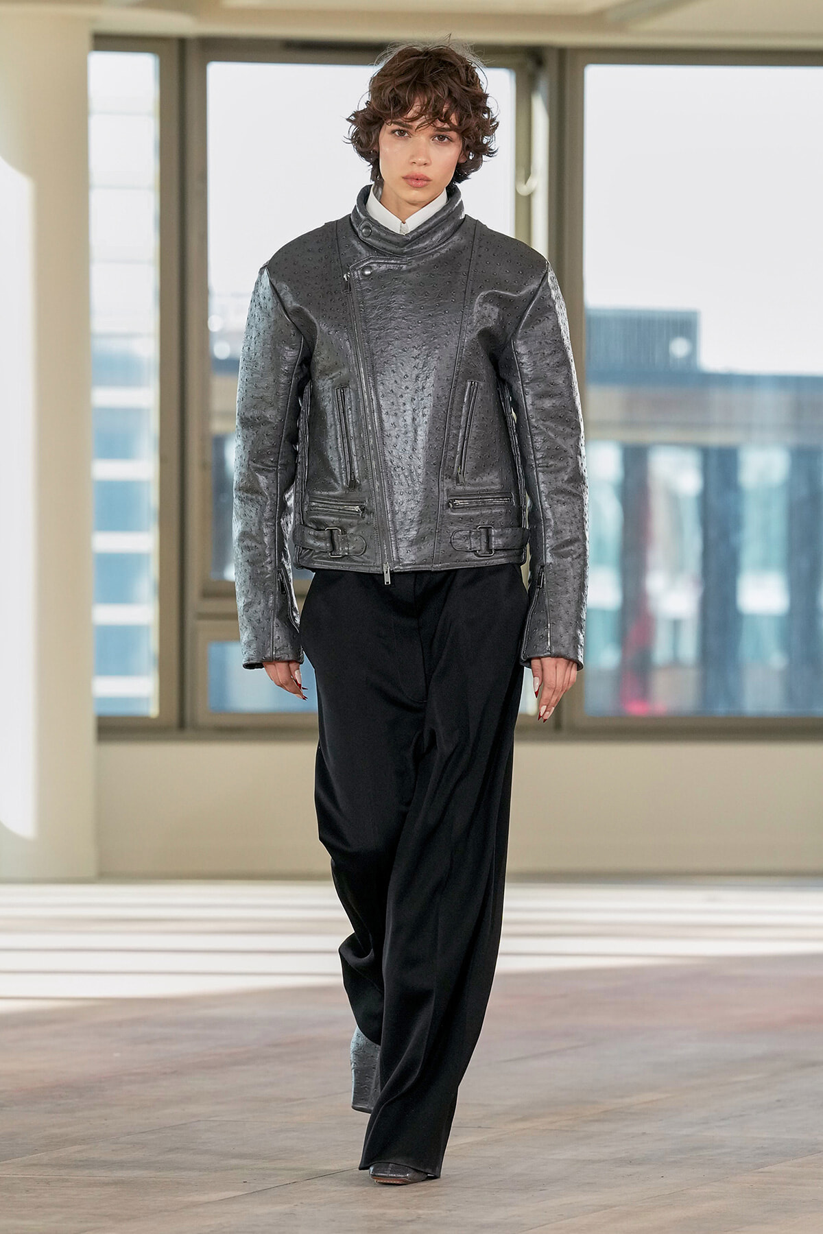 Model wearing a metallic gray textured biker jacket with zip pockets, paired with black wide-leg pants.