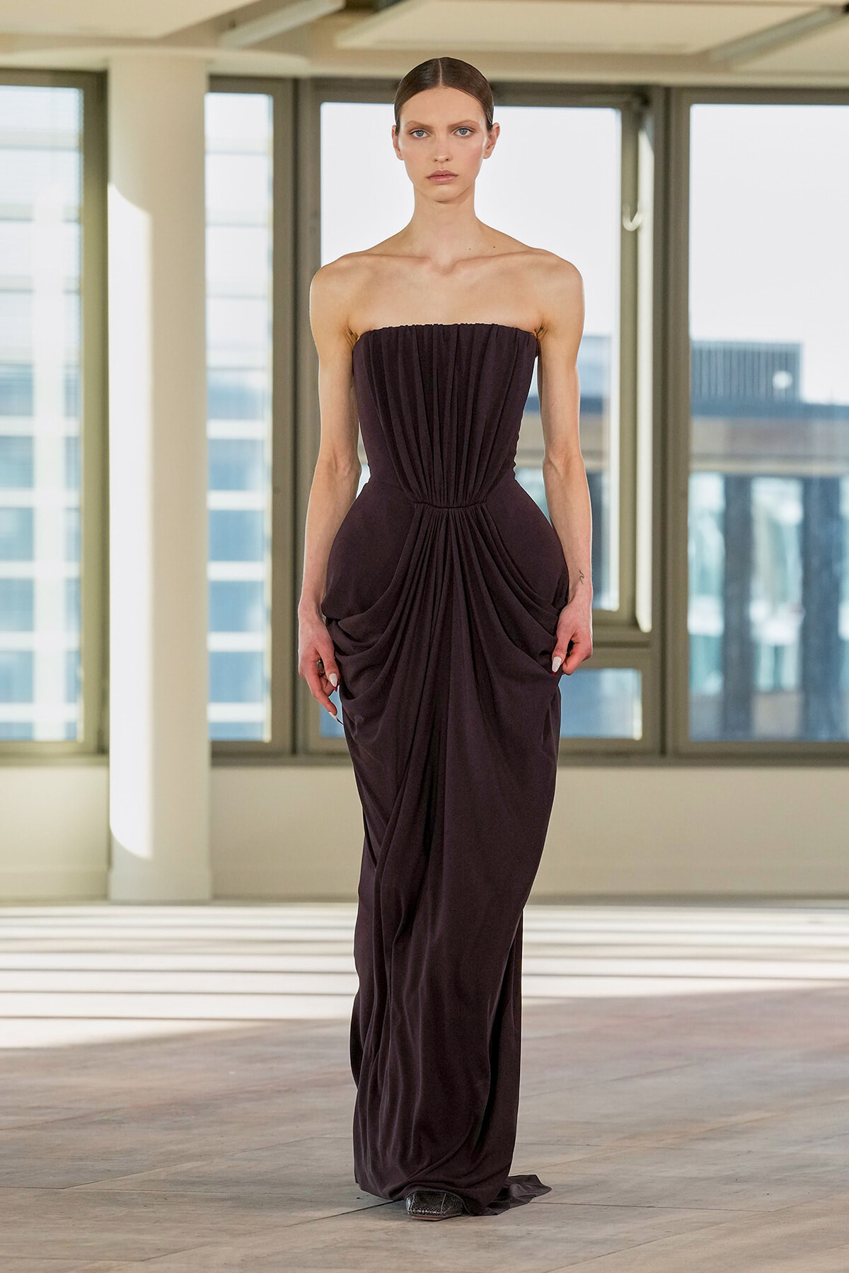 Model wearing a strapless, floor-length dark purple gown with gathered ruching at the bodice and draped folds along the skirt, standing in a bright studio with large windows