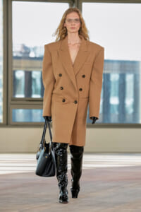 Model on a runway wearing a camel oversized double-breasted coat, black patent thigh-high boots, and a black handbag.