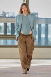Model walking the runway in a light blue oversized blazer over a matching cropped button-up bodysuit with midriff exposed, paired with tan loose-fit trousers and a brown belt, black heeled shoes.
