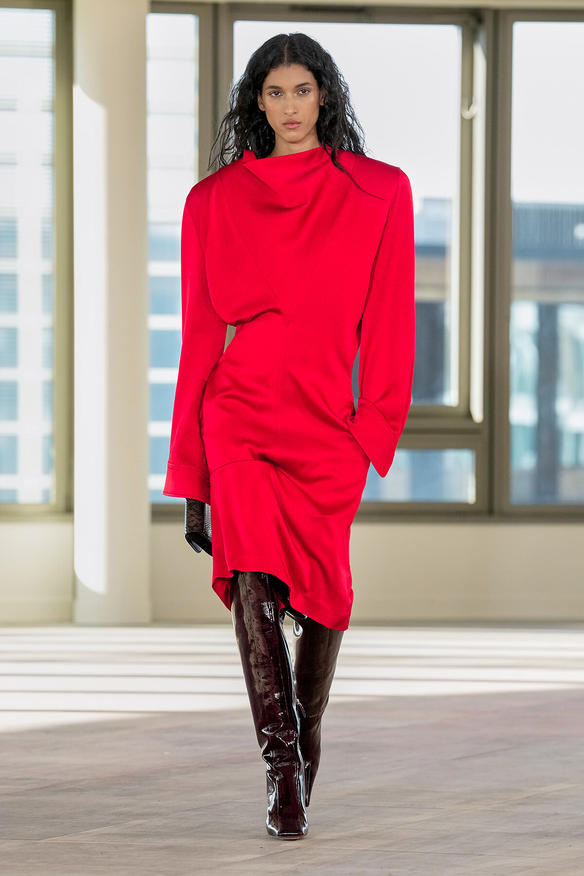 Female model in a bright red draped dress and dark patent thigh-high boots walking indoors.