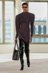 Model wearing a dark purple ruched dress with side panels, black patent thigh-high boots, sunglasses, burgundy gloves, and a textured handbag.