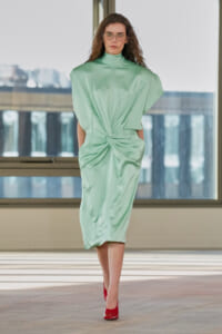 Model walking a runway in a mint green draped midi dress with a twisted waist, paired with red heels in a bright, modern venue.