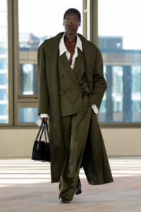 Model wearing an oversized olive trench coat over a double-breasted suit with a white shirt, carrying a black handbag while walking indoors in a fashion setting.