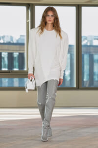 Model on a runway wearing an oversized white top with draped sides, glossy gray patent pants, and a white handbag.