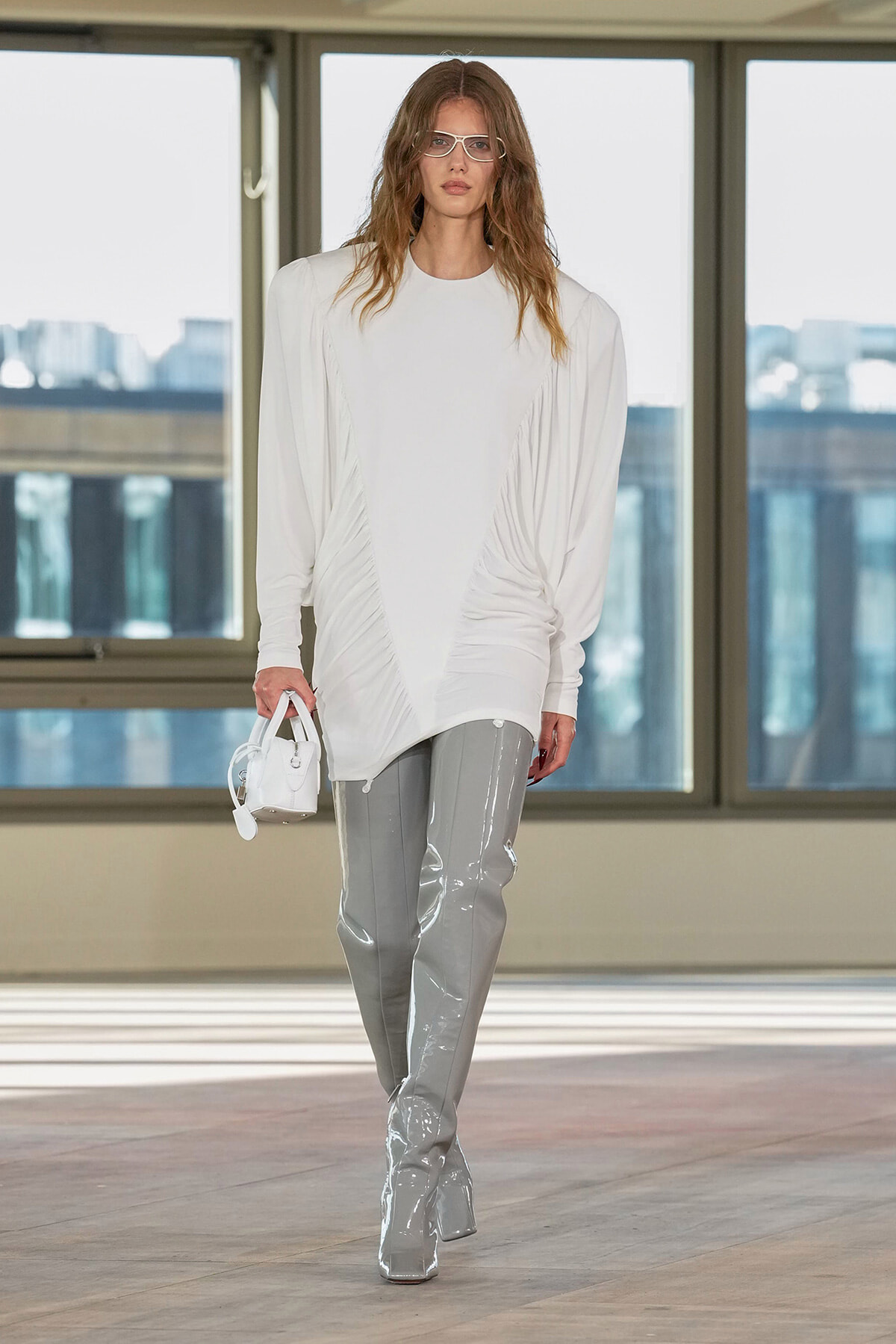 Model on a runway wearing an oversized white top with draped sides, glossy gray patent pants, and a white handbag.