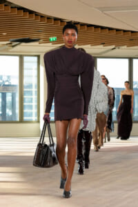 Model walks a runway in a dark plum mini dress with ruched sides, wearing gloves and carrying a black textured handbag.