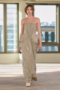 Model wearing a strapless, floor-length, glittering champagne gown walking down a runway indoors.