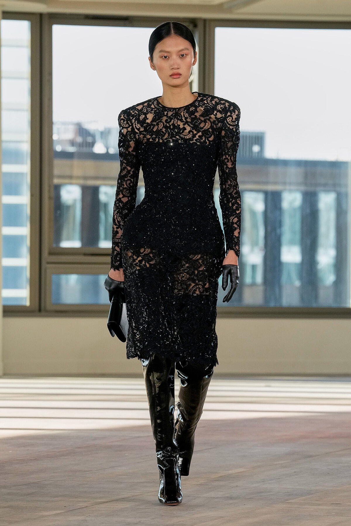 Model on a runway wearing a fitted black lace evening dress with sheer sleeves, sparkling embellishments, black gloves, and glossy knee-high boots.