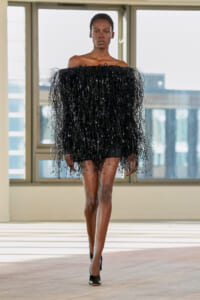 Model wearing a black off-the-shoulder mini dress with long fringe walking toward the camera on a runway in a bright studio.