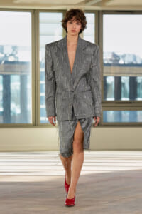 Model walking in a grey pinstripe suit with fringe embellishments, deep V-neck, matching skirt with a high slit, red patent heels in a studio setting.