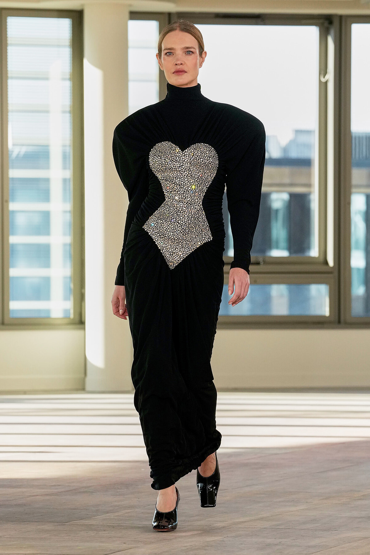 Model wearing a black velvet gown with a large rhinestone heart applique at the torso, walking in a bright studio space with windows behind.