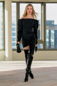 Model walking in a black off-shoulder mini dress with shiny thigh-high boots, carrying a small black clutch in a sunlit indoor space.