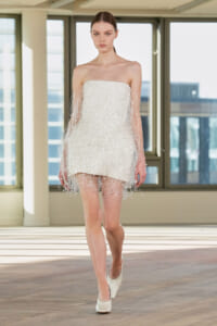 Model walks in a strapless white beaded mini dress with fringe beads on a runway indoors during a fashion show