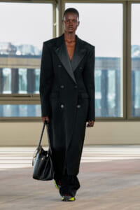 Model in a long black double-breasted coat, holding a black handbag, walking in a bright studio with large windows.